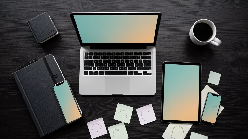Overhead view of a laptop, tablet, and phone on a dark wood desk with a notebook, coffee cup, and paper notes